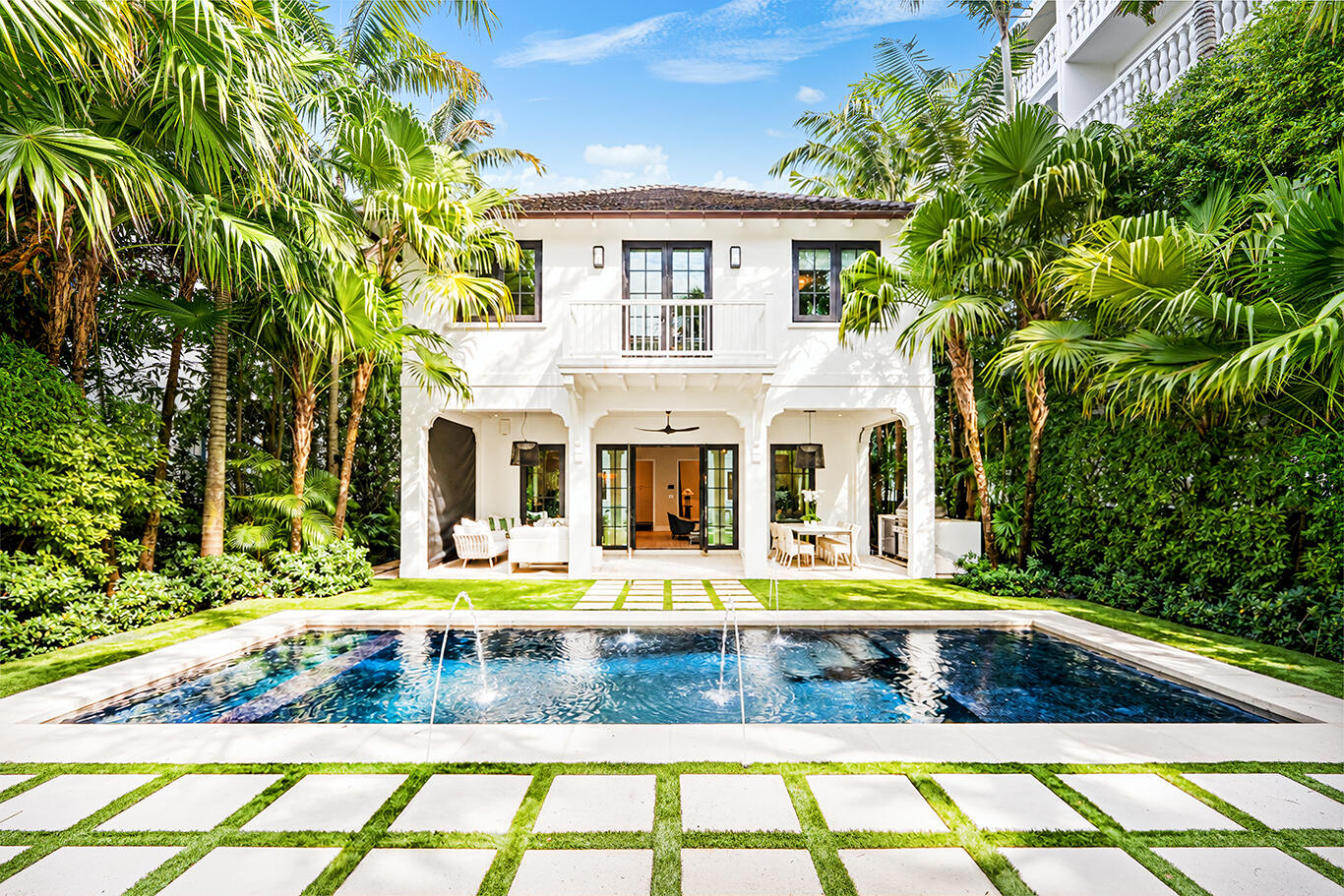 White two-story home behind a swimming pool, framed by palms and tropical landscaping.