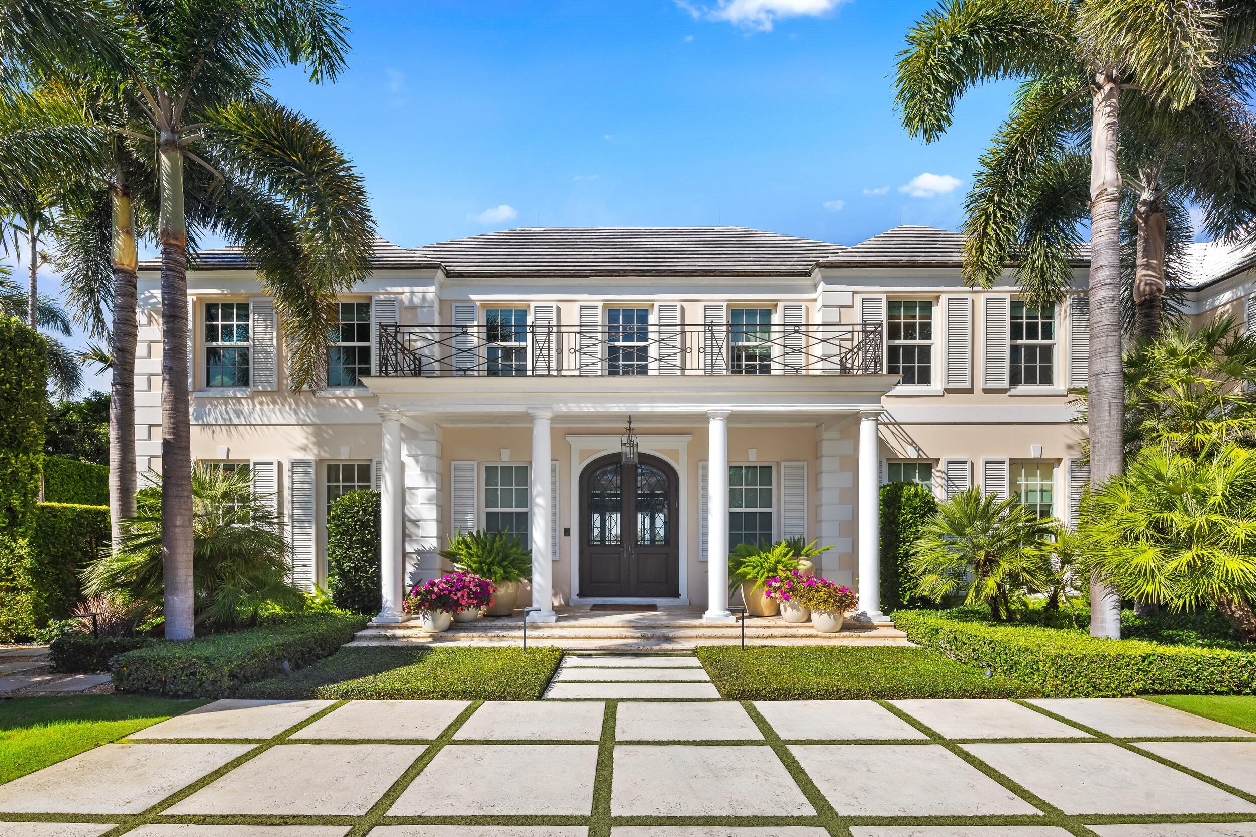 Two-story residence with a columned entrance, balcony, palms, and broad stone walkway.