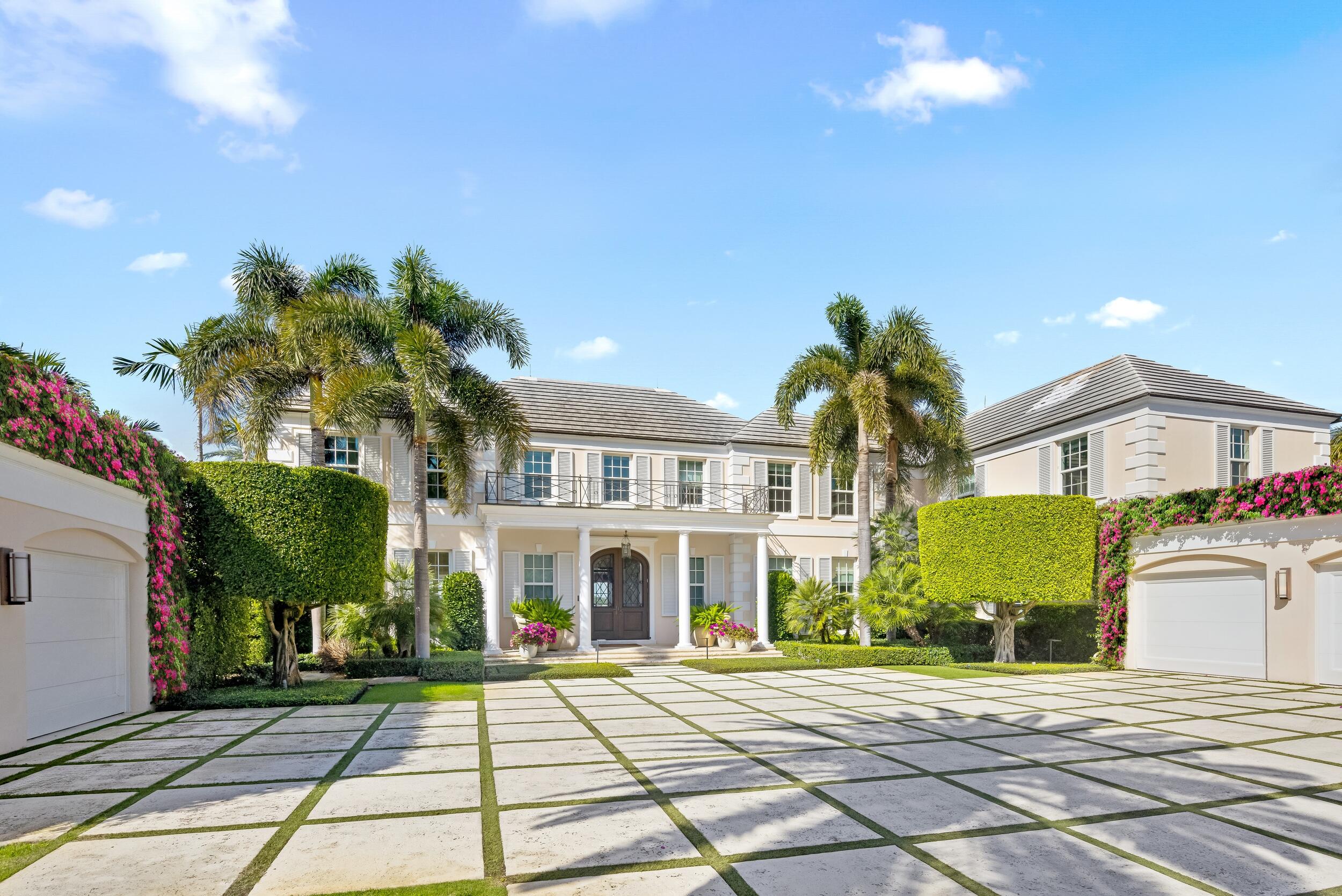 Two-story cream home with a columned entrance, palm trees, flowering vines, and a broad paved driveway.