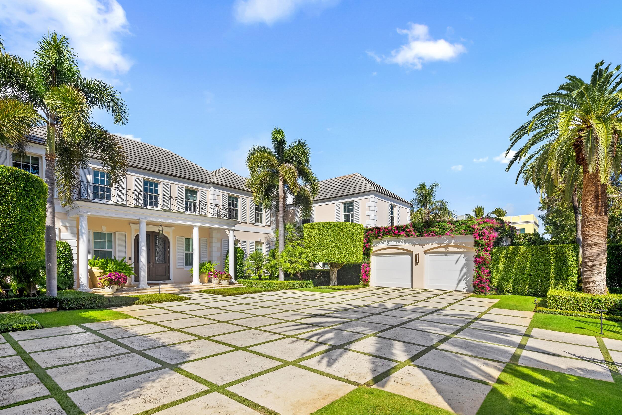 Two-story residence with a columned porch, bougainvillea-framed garage, palms, and a stone-and-grass driveway.