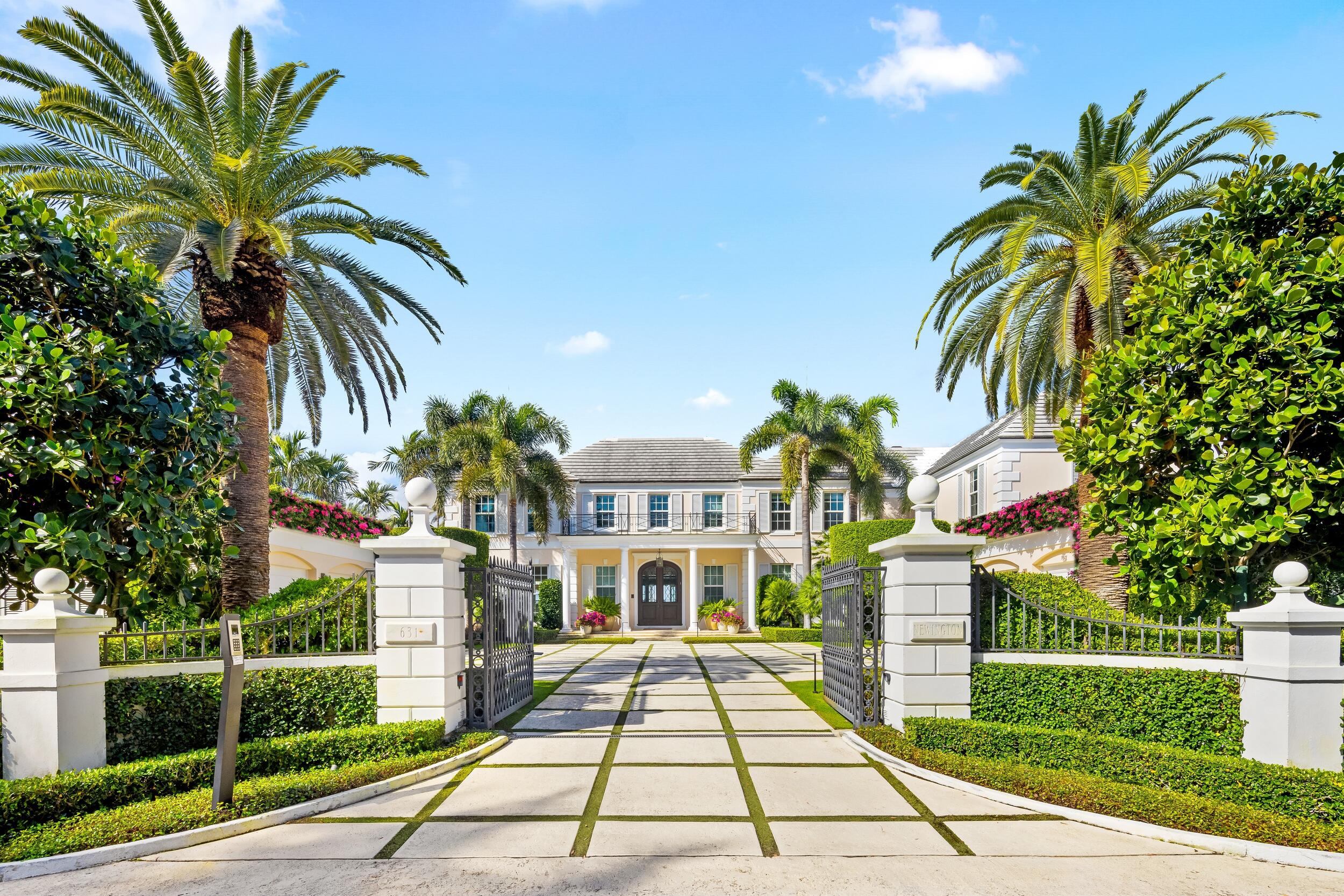 White two-story residence behind open gates, with a geometric driveway, tall palms, and lush landscaping.