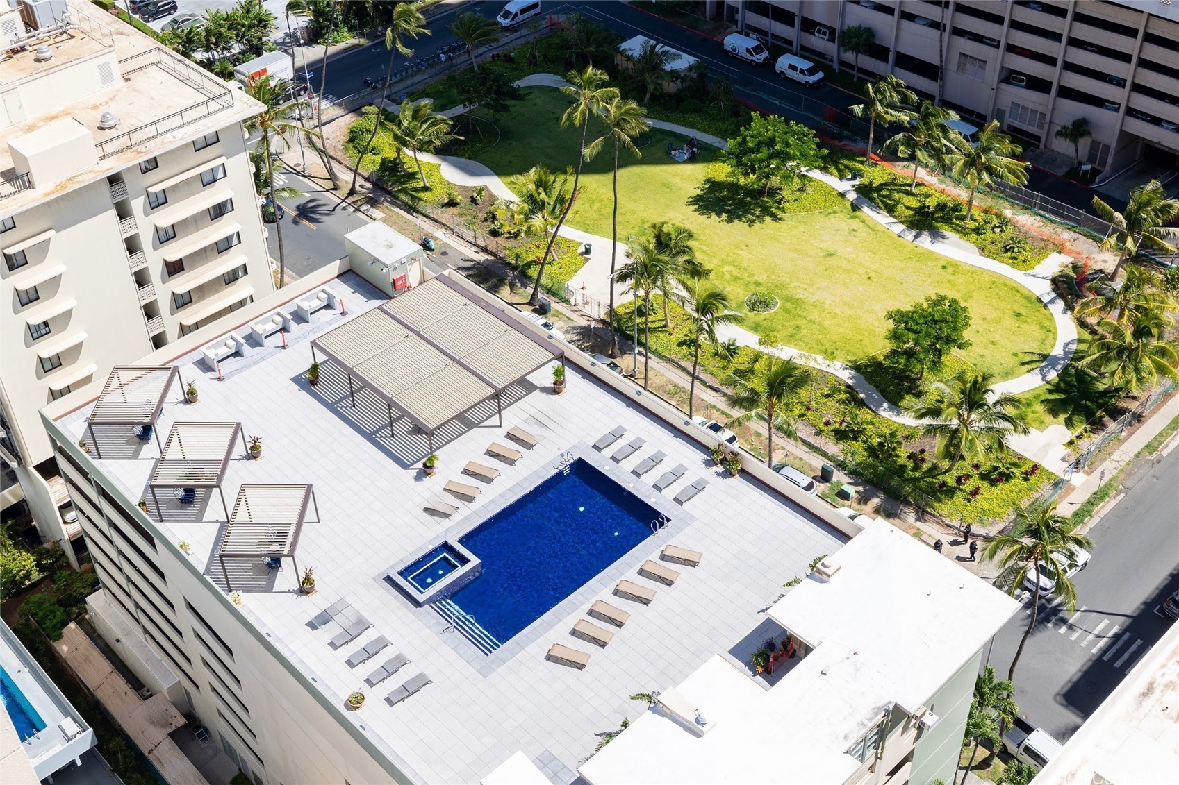 Aerial view of a condominium pool deck with a blue swimming pool, shaded seating, and a palm-lined lawn.