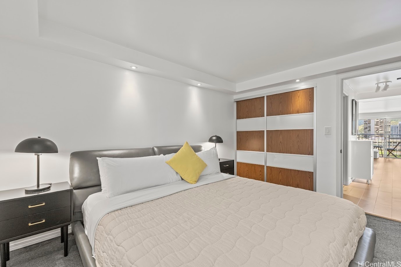 Staged bedroom with a gray upholstered bed, wood-paneled closet doors, and a view toward the living area.
