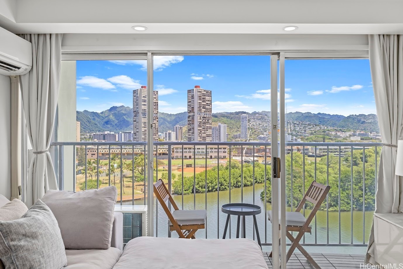 Bedroom with balcony seating overlooking a canal, Honolulu high-rises, and green mountains.