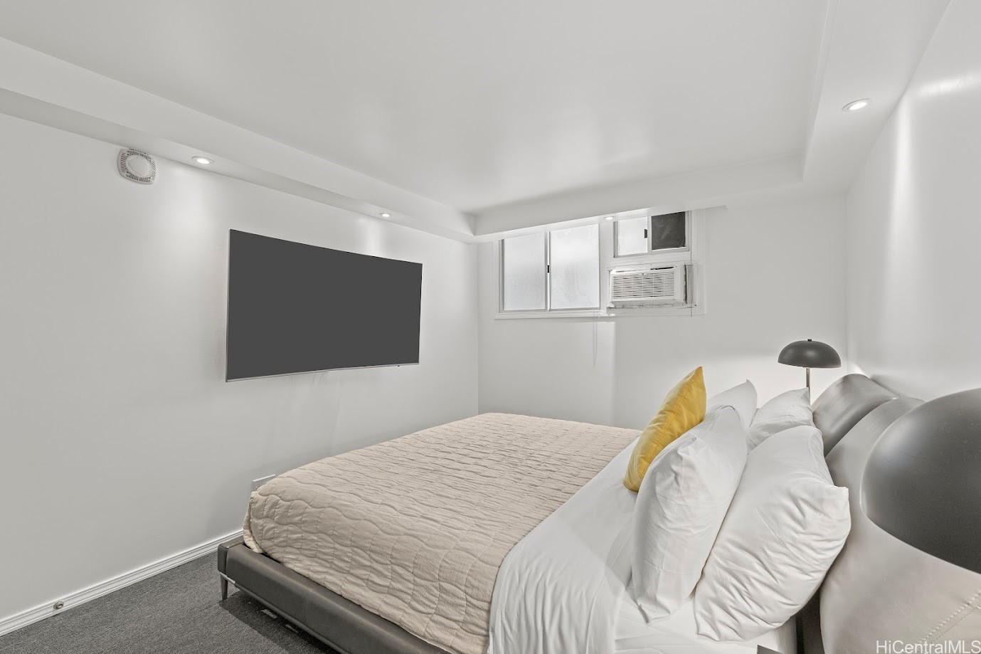 Bedroom with a quilted bed, white pillows, wall-mounted television, and window air conditioner.
