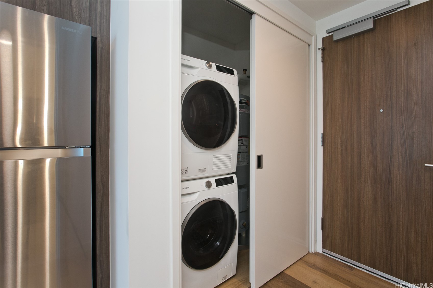 Laundry closet with stacked Samsung washer and dryer beside a stainless-steel refrigerator.