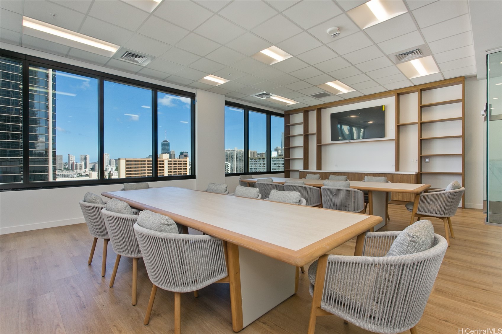 Conference room with long tables, gray chairs, built-in shelving, and city views through large windows.