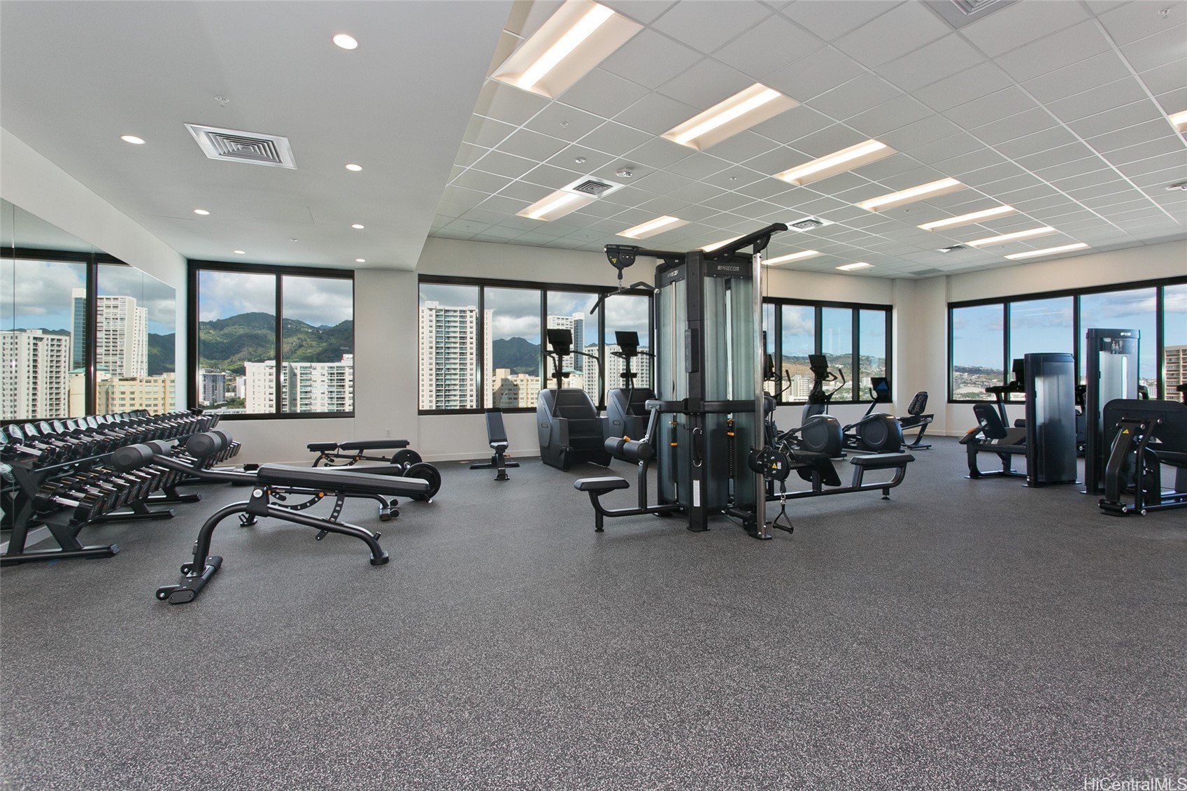 Fitness center with weight racks and cardio machines, overlooking Honolulu high-rises and mountains.
