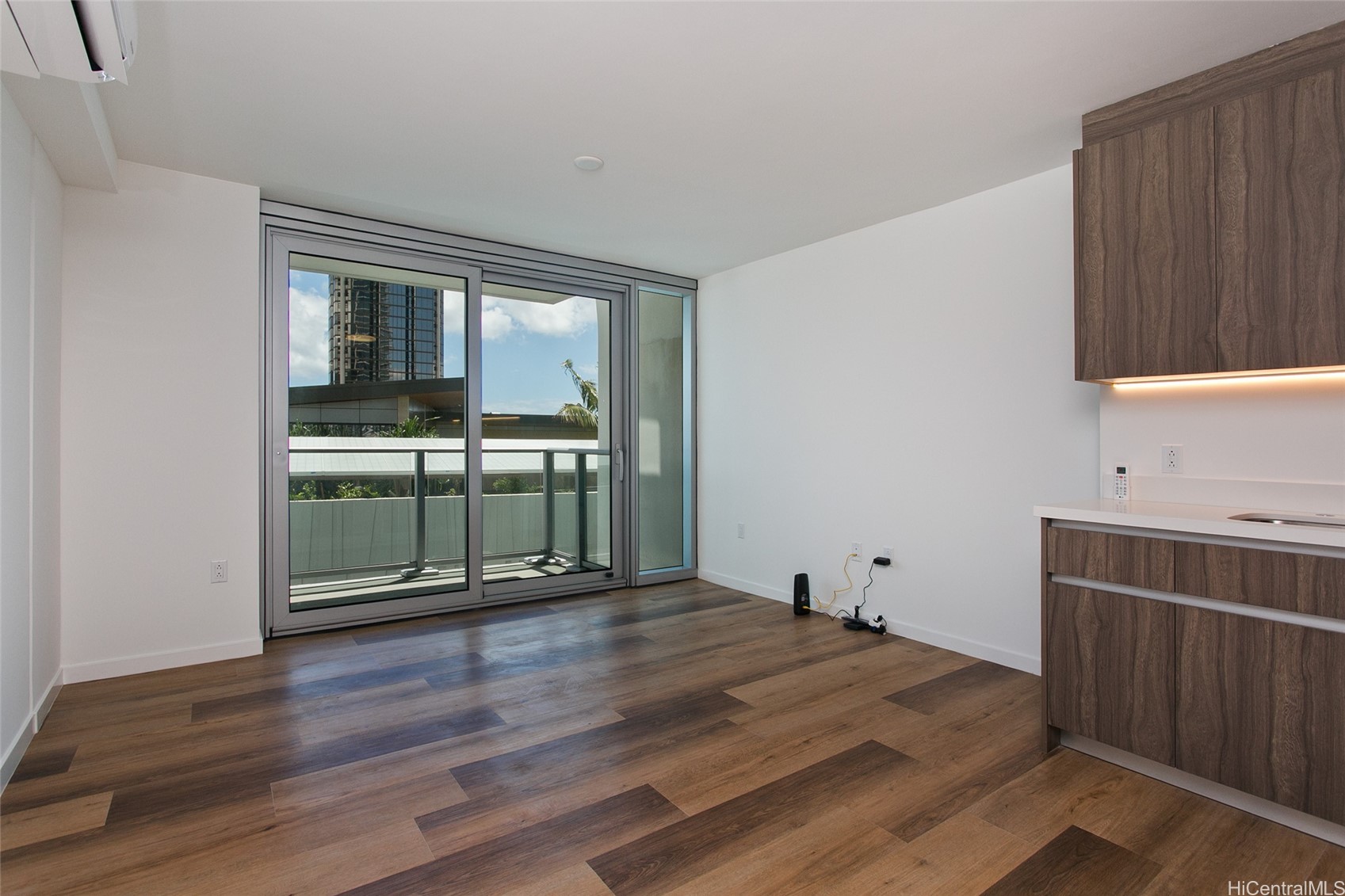 Unfurnished condo living area with wood-look floors, dark kitchen cabinets, and sliding balcony doors.