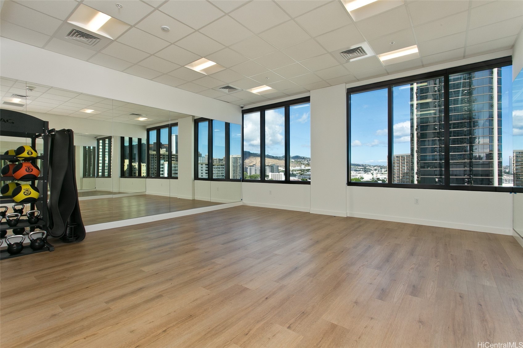 Exercise room with mirrored wall, wood-look floors, and large windows overlooking Honolulu high-rises.