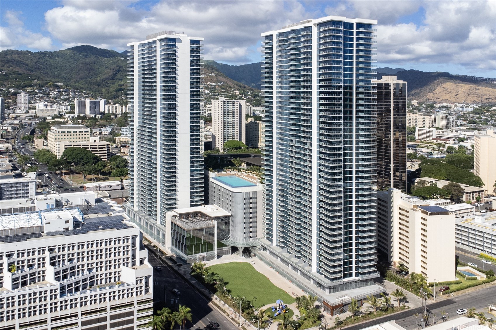 Two modern residential towers with a pool, Honolulu city buildings, and green mountains behind them.