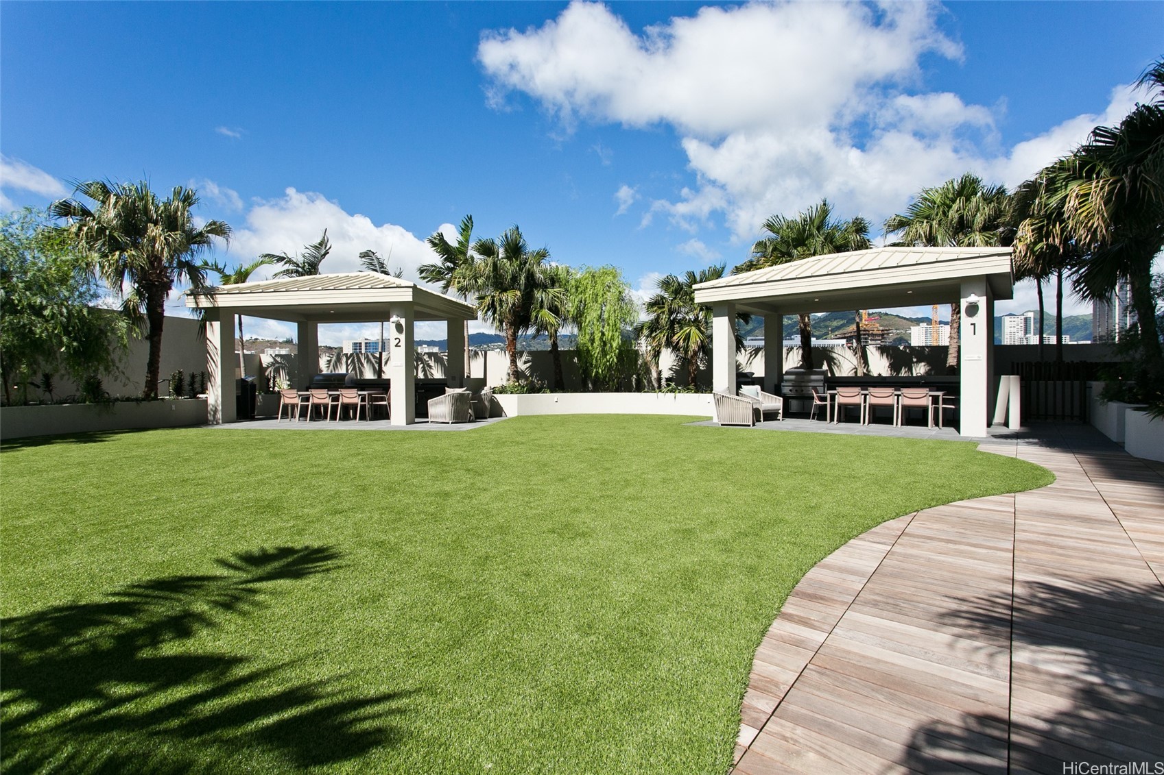 Outdoor amenity terrace with two covered barbecue pavilions, a lawn, palm trees, and distant mountains.