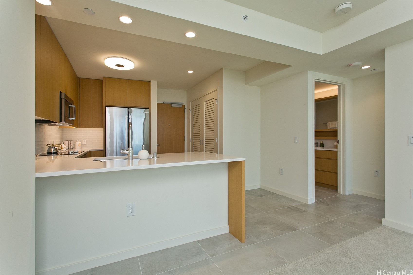 Vacant condo kitchen with light wood cabinets, stainless steel refrigerator, and a white peninsula counter.