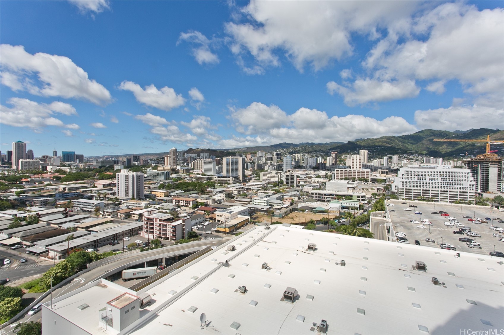 Elevated Honolulu view of the city skyline and green mountain ridges beneath a partly cloudy sky.