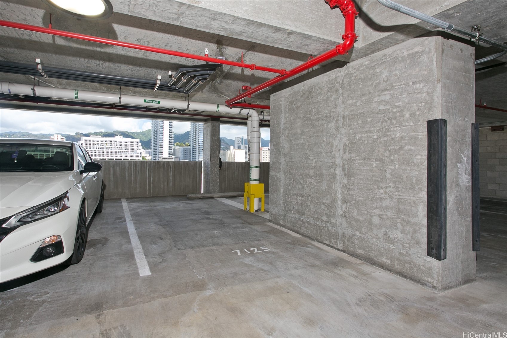 Concrete parking garage stall 7125 with exposed red pipes and Honolulu buildings visible outside.