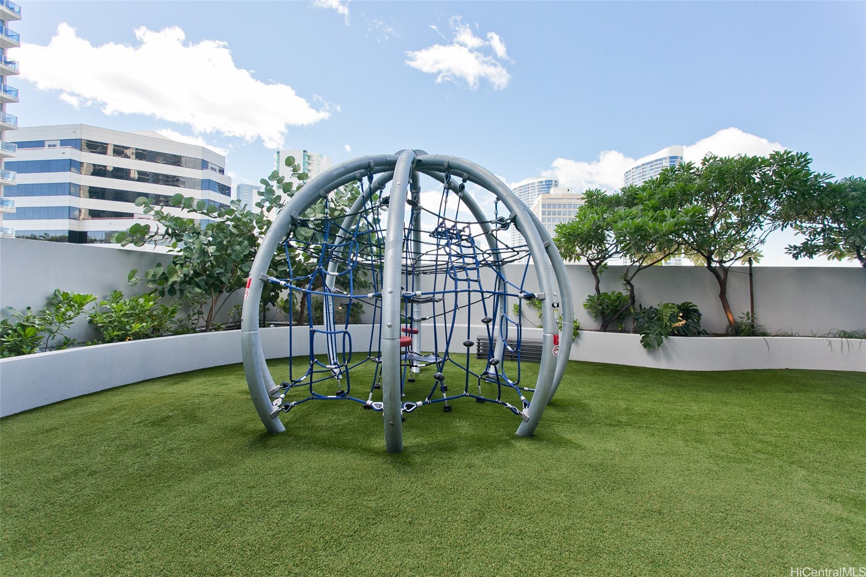 Rooftop playground with a metal and rope climbing structure, artificial turf, and landscaped planters.