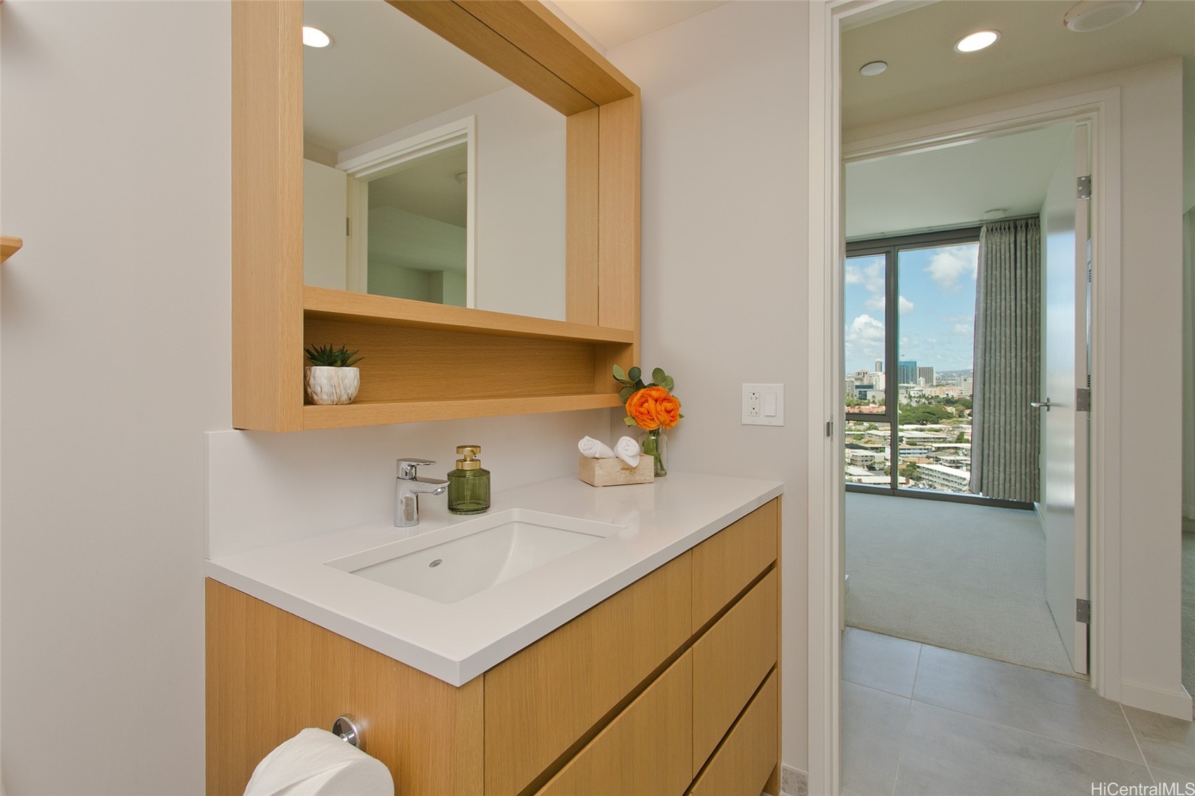 Bathroom vanity with light wood cabinetry and a bedroom window overlooking Honolulu buildings