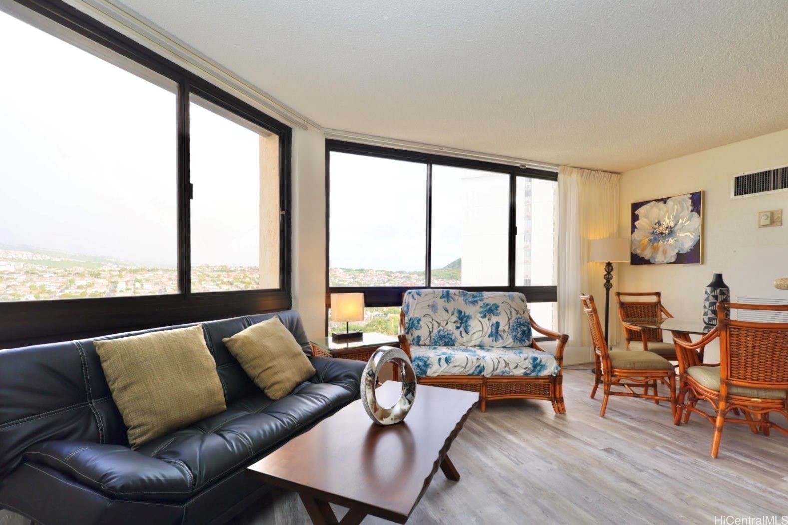 Furnished living room with corner windows overlooking a neighborhood and distant green hills.