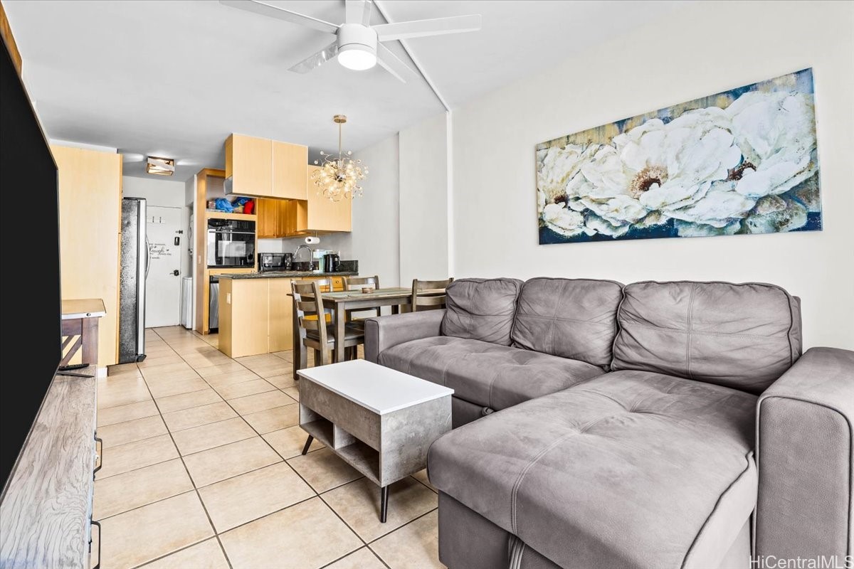 Open-plan living area with a gray sectional, tiled floors, and light wood kitchen cabinetry.
