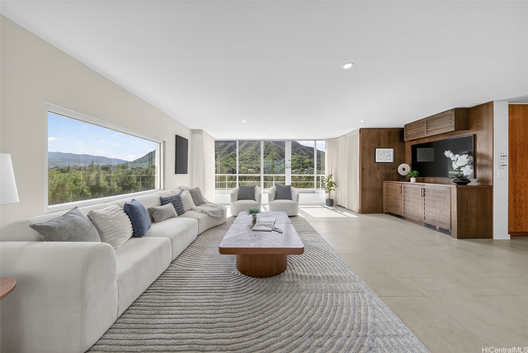 Living room with a white sectional, wood media wall, and broad windows overlooking green mountains.