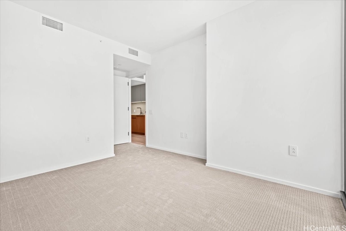 Empty carpeted room with white walls and an open doorway to the kitchen.