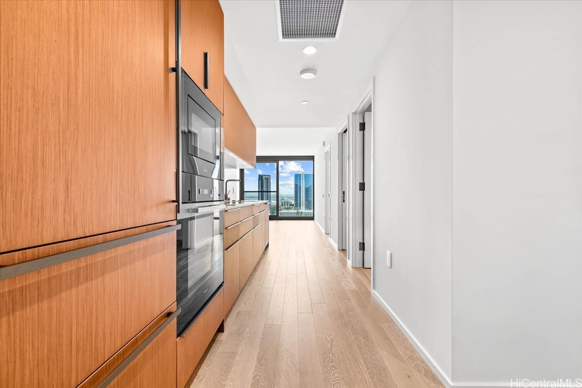 Modern kitchen hallway with warm wood cabinetry, built-in appliances, and a city view through the far window.