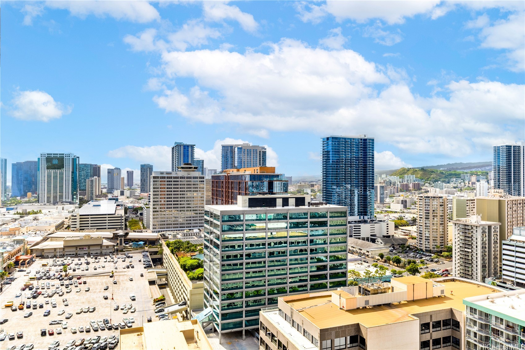 Honolulu skyline with high-rise buildings, a foreground parking lot, and green hills in the distance.