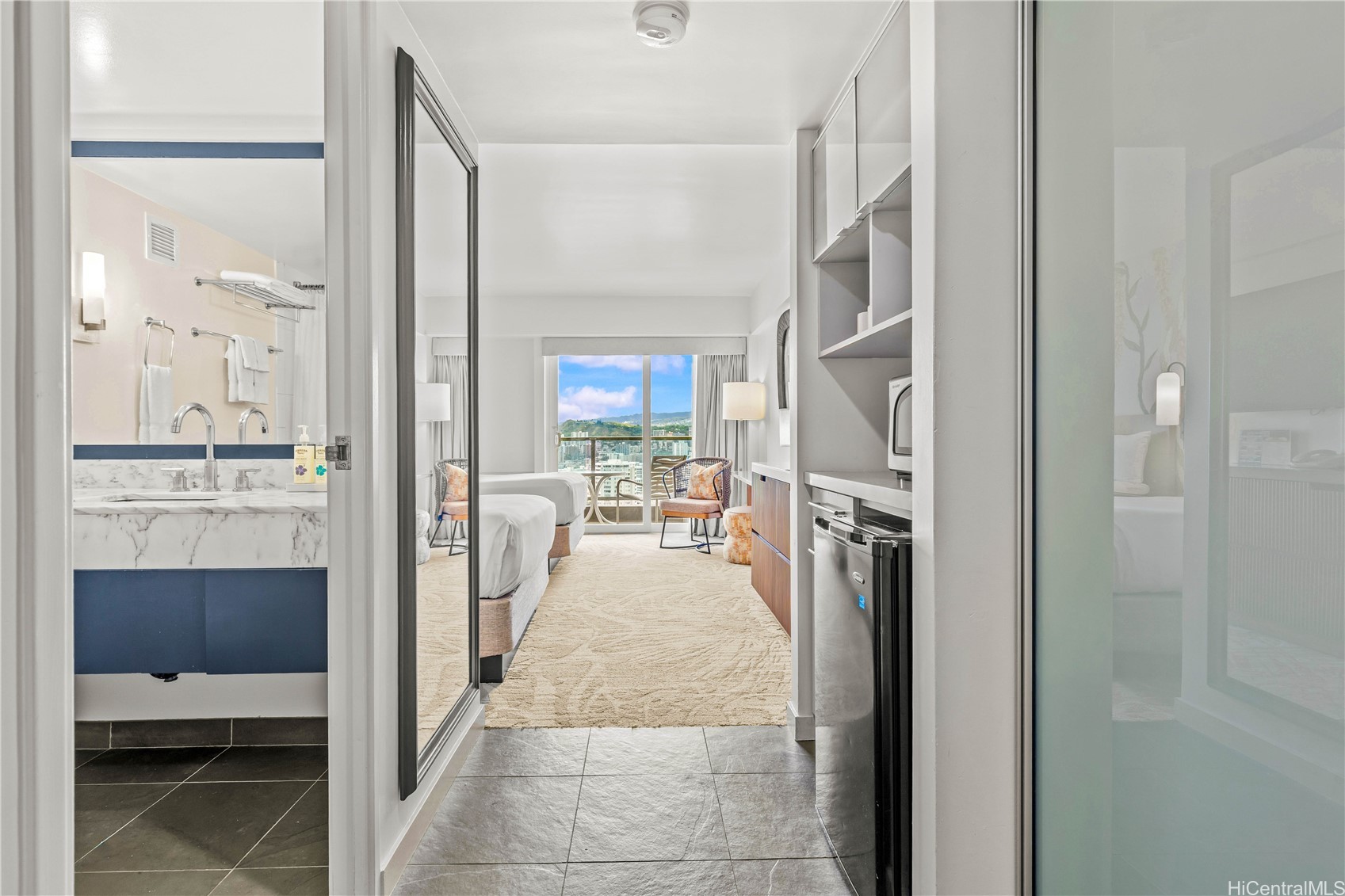 Furnished studio with marble bathroom vanity, kitchenette, and balcony view of Honolulu buildings and mountains.