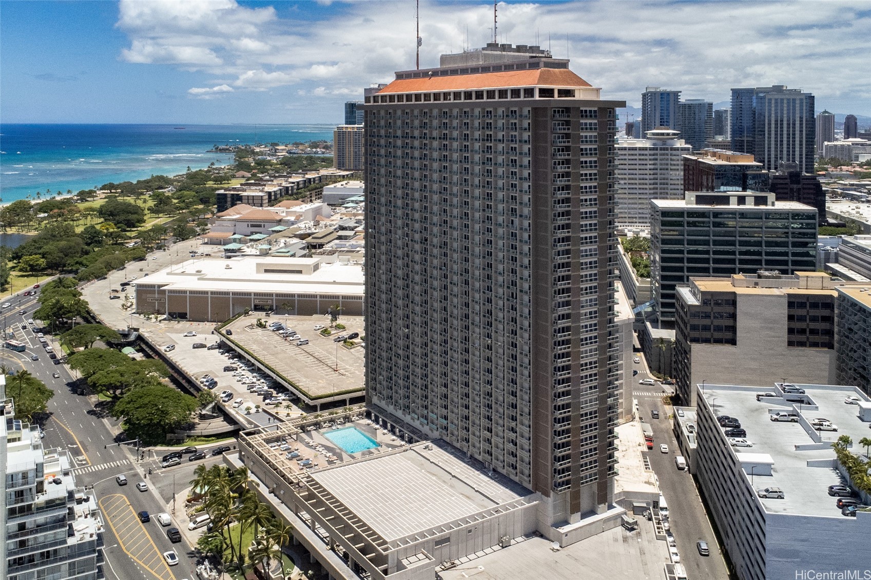 Honolulu condominium tower beside the ocean, viewed from above with a pool on its lower deck.