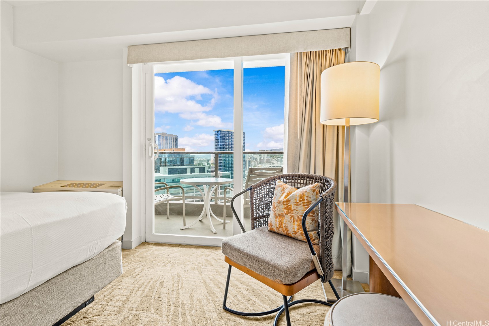 Furnished bedroom with a woven lounge chair and balcony overlooking Honolulu high-rises.