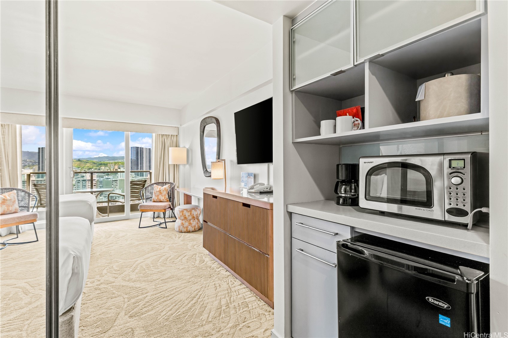 Furnished studio with a compact kitchenette and balcony overlooking city buildings and a green mountain ridge.