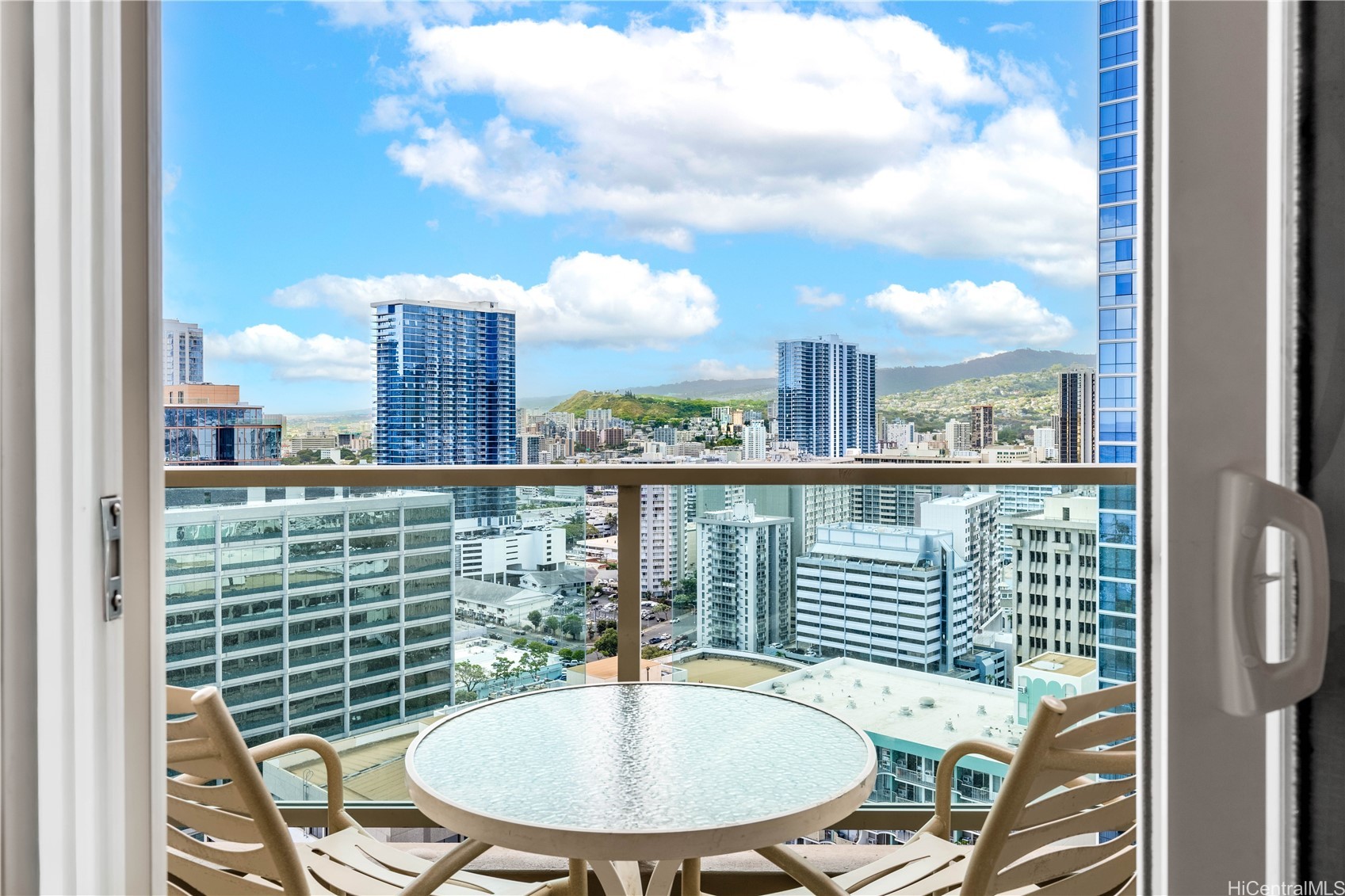 High-rise balcony with a glass-top table and chairs overlooking Honolulu’s cityscape and green hills.
