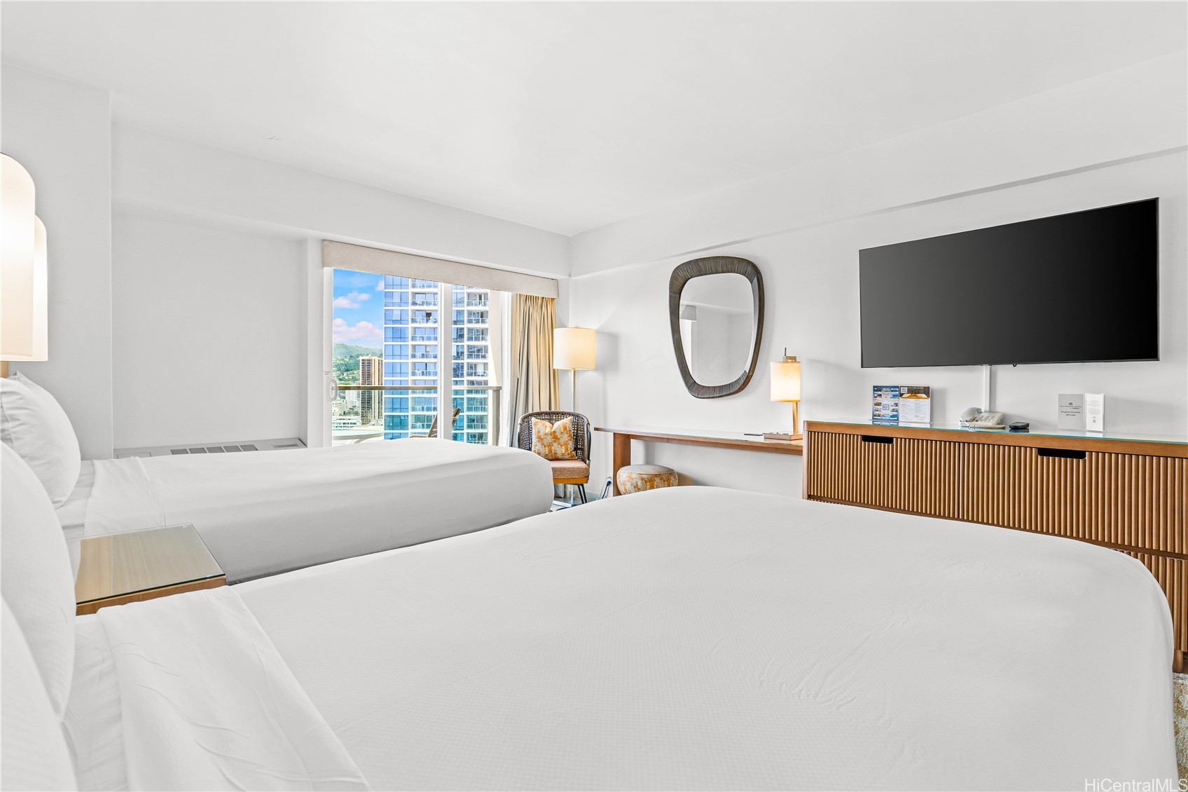 Furnished bedroom with two white beds and sliding doors overlooking high-rise buildings.