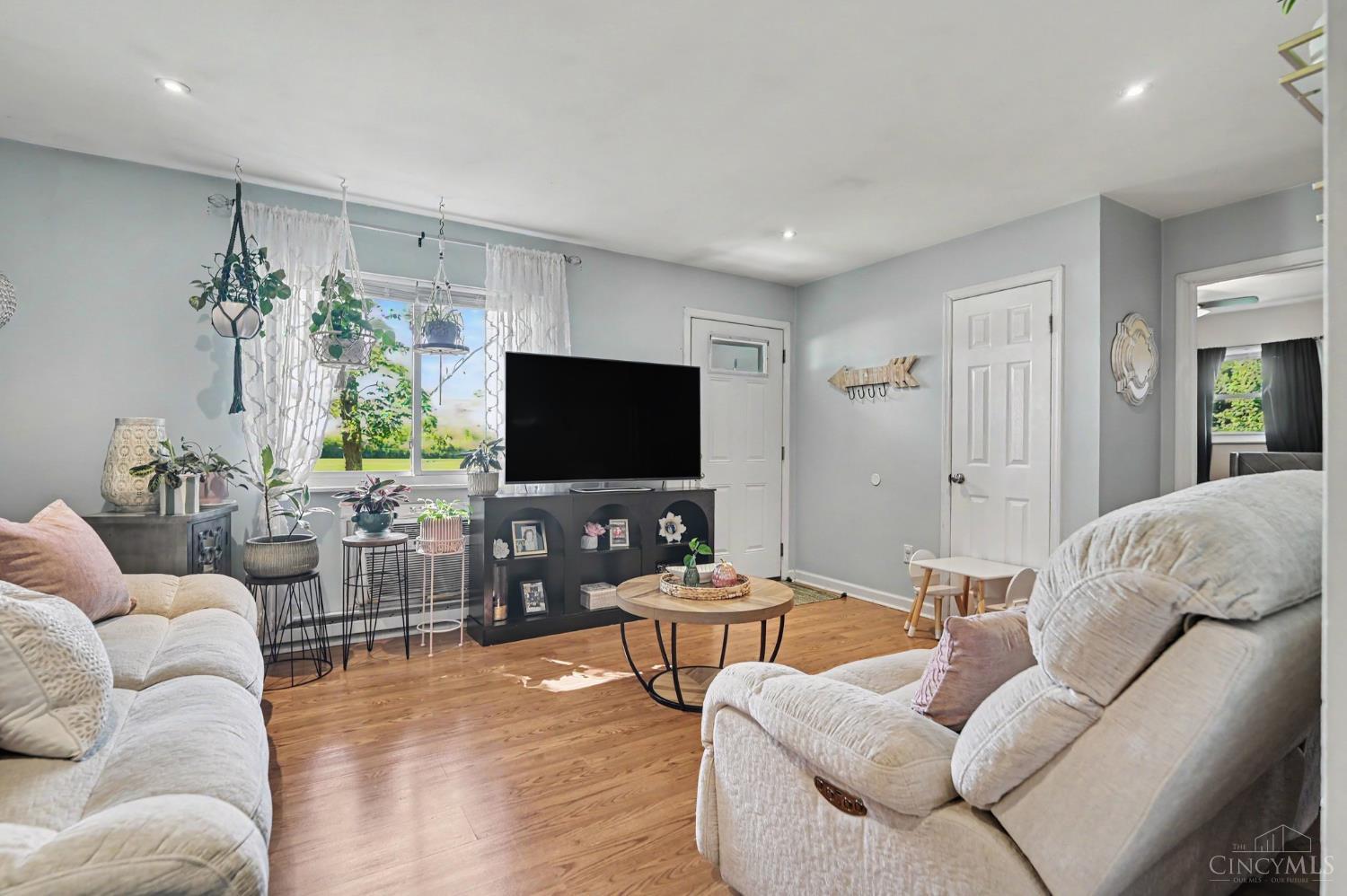 Living room with hardwood floors, pale sofas, a television, and hanging plants by a large window.