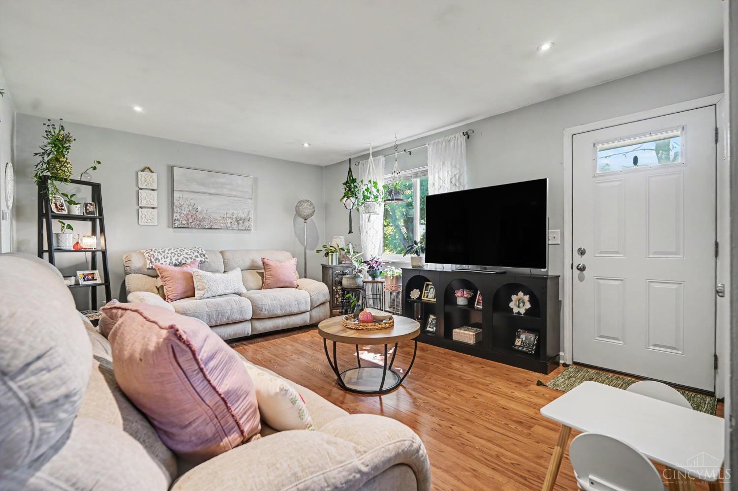 Living room with cream sofas, blush pillows, hardwood floors, and a large television.