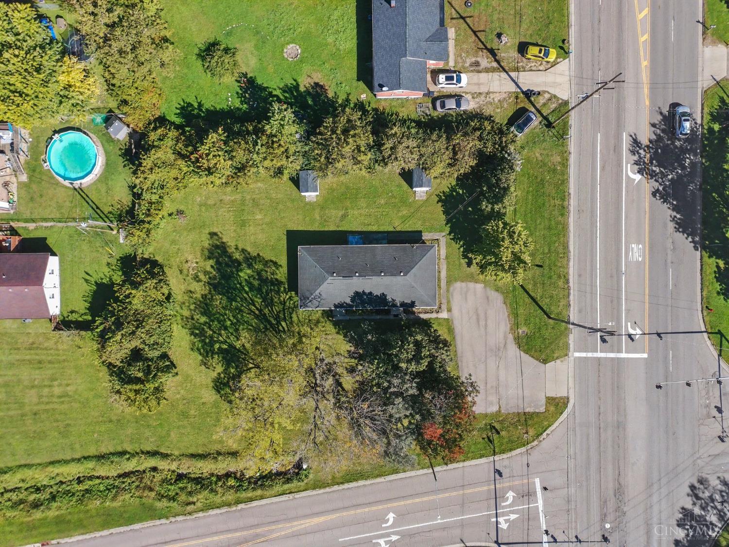 Aerial view of a roadside property with a lawn, mature trees, and a nearby pool.