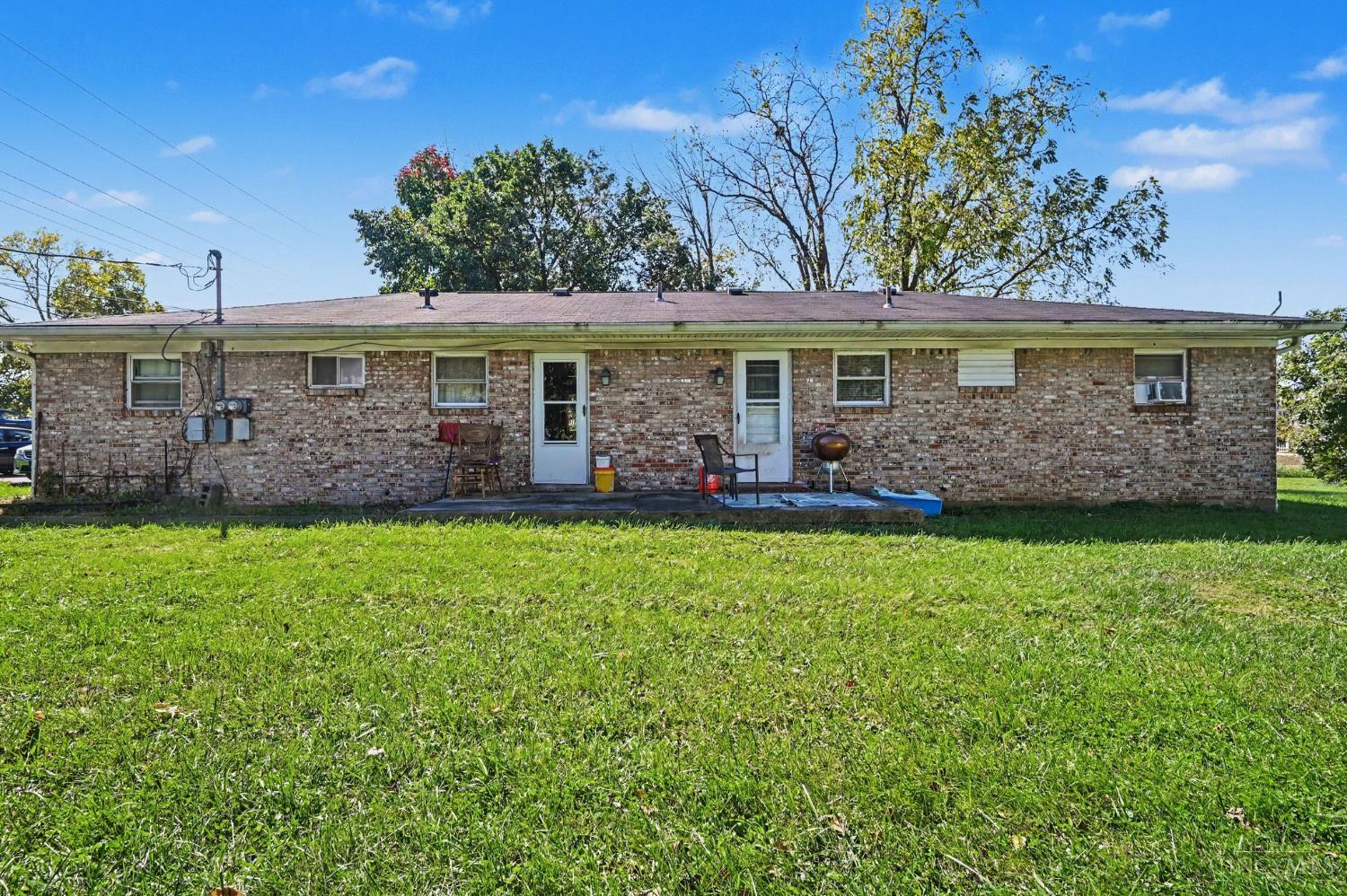 Rear of a single-story brick building with patio chairs, a grill, and a broad lawn.