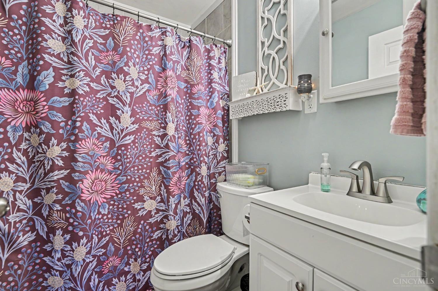 Bathroom with a white vanity, toilet, pale blue walls, and a bold purple floral shower curtain.