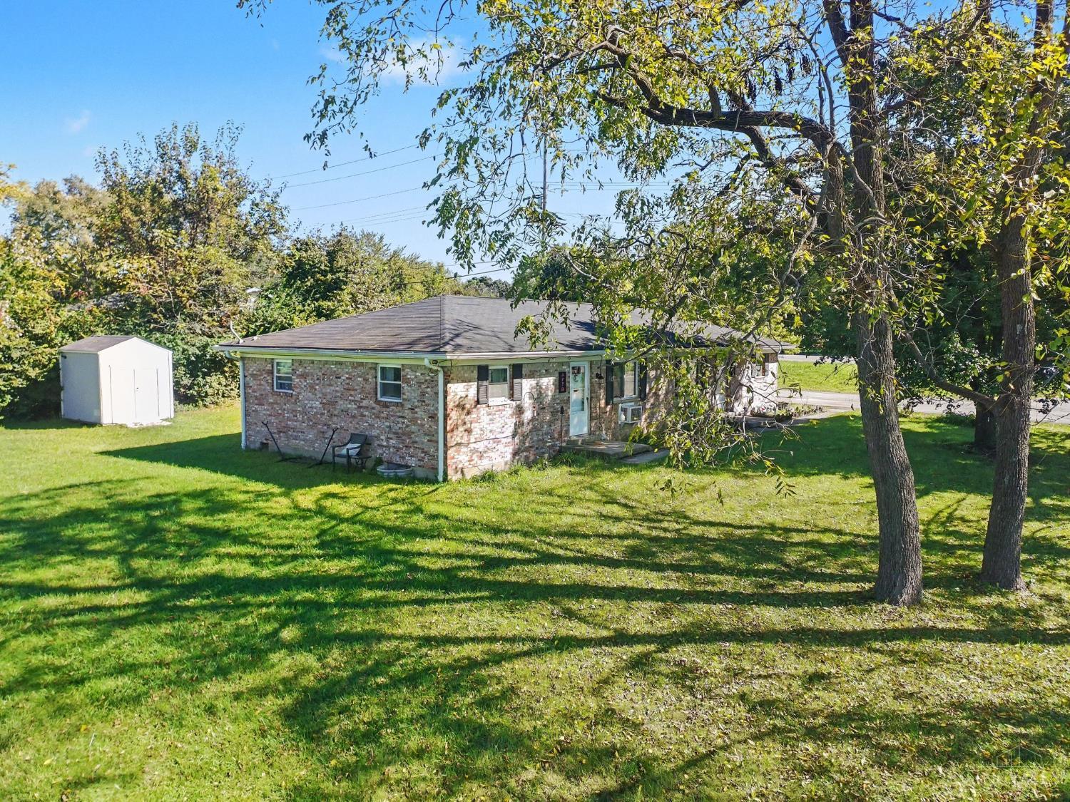 Single-story brick house with a white storage shed and mature trees across a grassy yard.