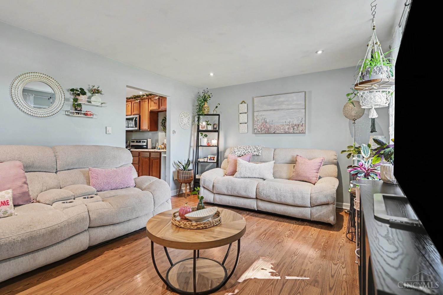 Furnished living room with two light sofas, pink pillows, hardwood floors, and an open kitchen doorway.