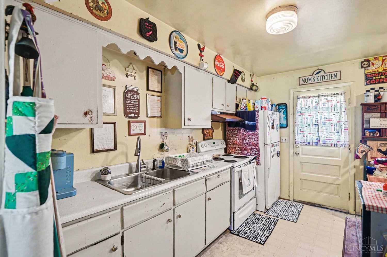 Kitchen with worn white cabinets, a double sink, electric range, and a door labeled “Mom’s Kitchen.”