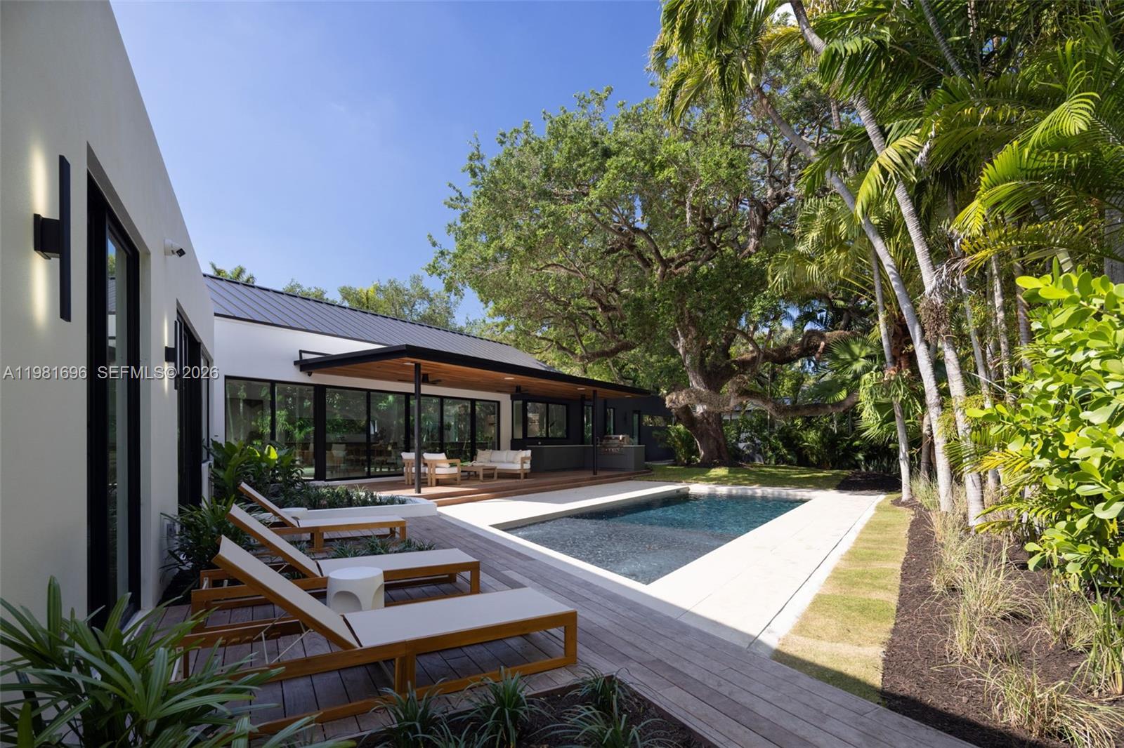 Modern home with a rectangular pool, wood deck, and tropical landscaping beneath a large oak tree.