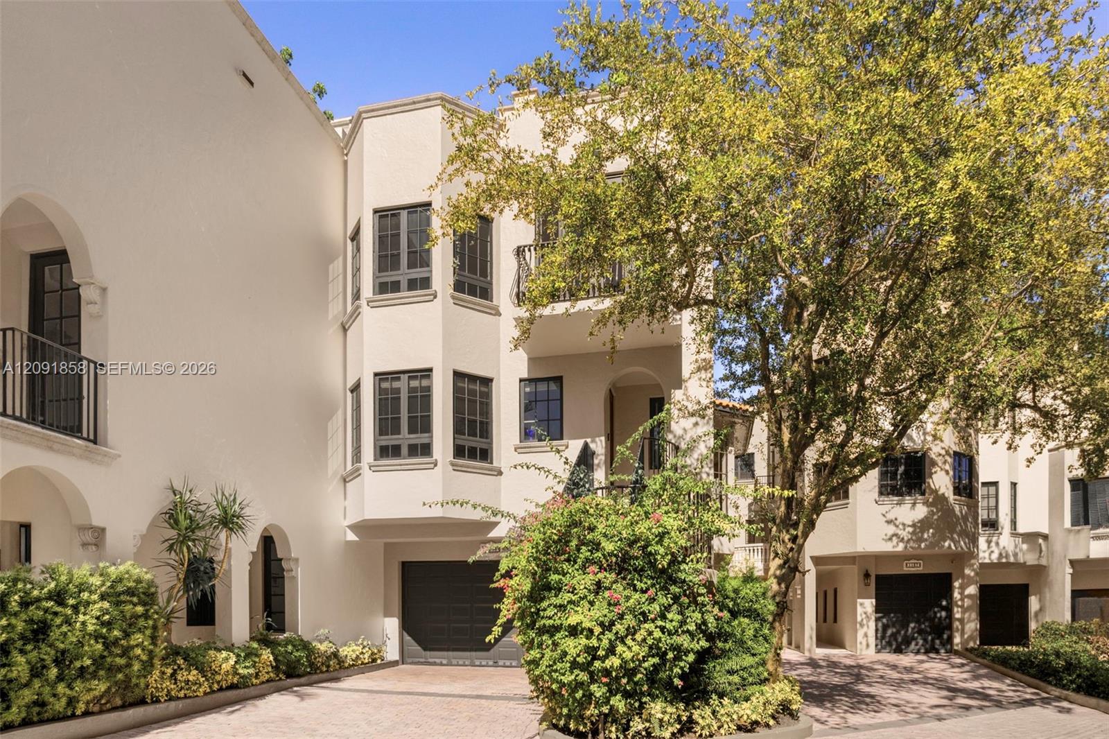 Cream stucco townhouses with arched entrances and balconies beneath a large leafy tree.