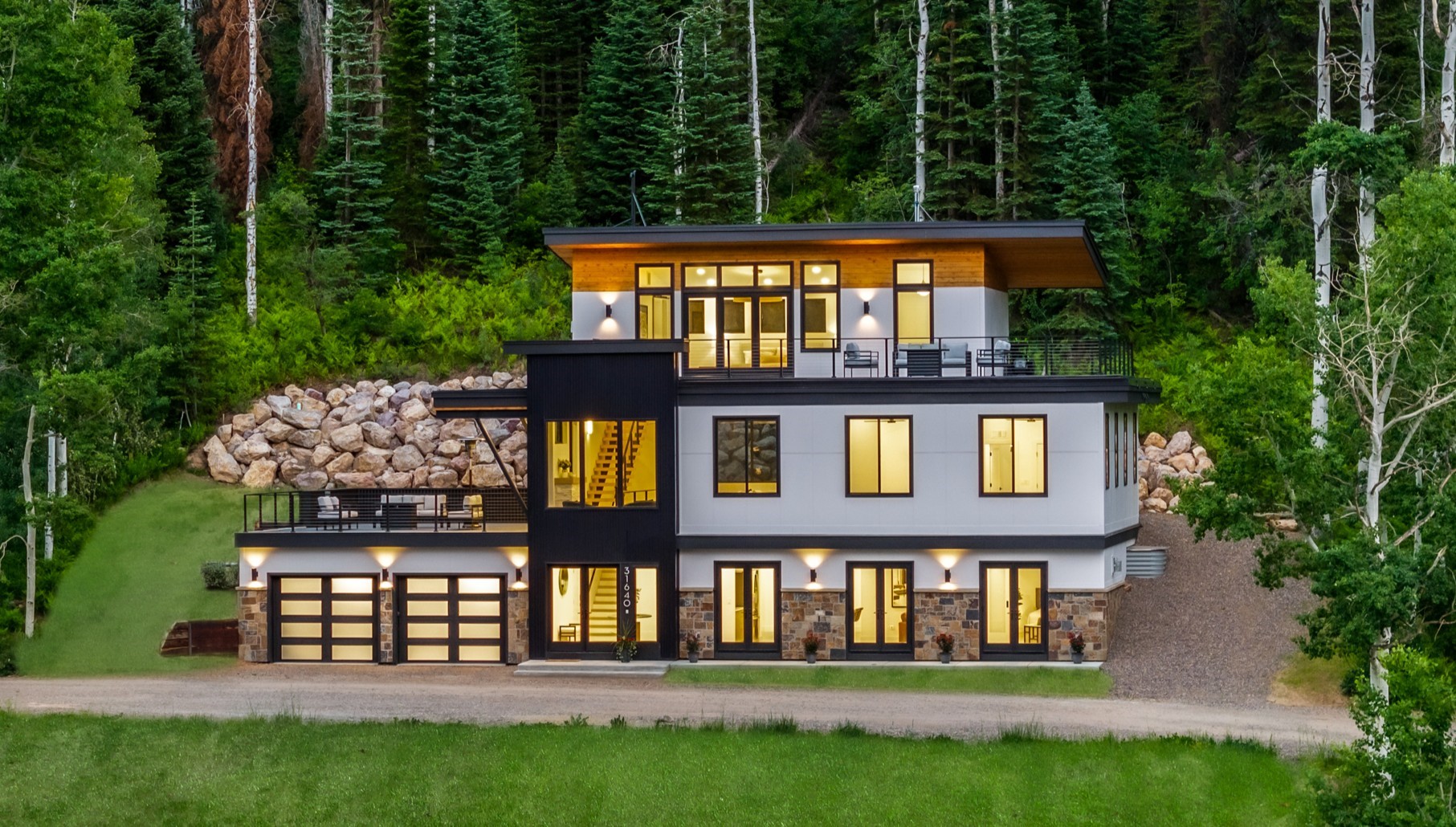 Modern multi-level home with lit windows, broad decks, and a forested setting at dusk.