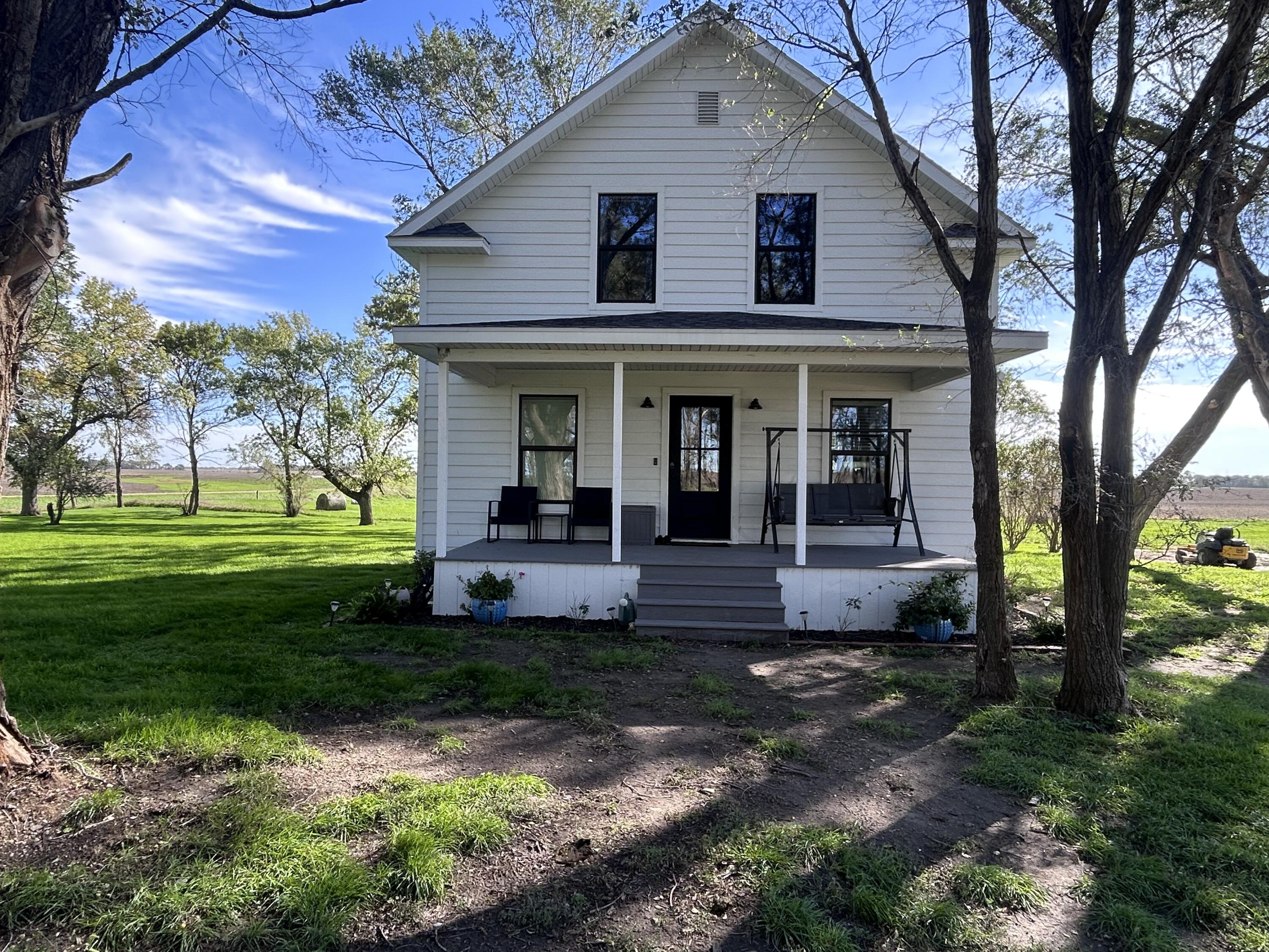 White two-story farmhouse with a covered porch and swing beneath mature trees on a green lawn.