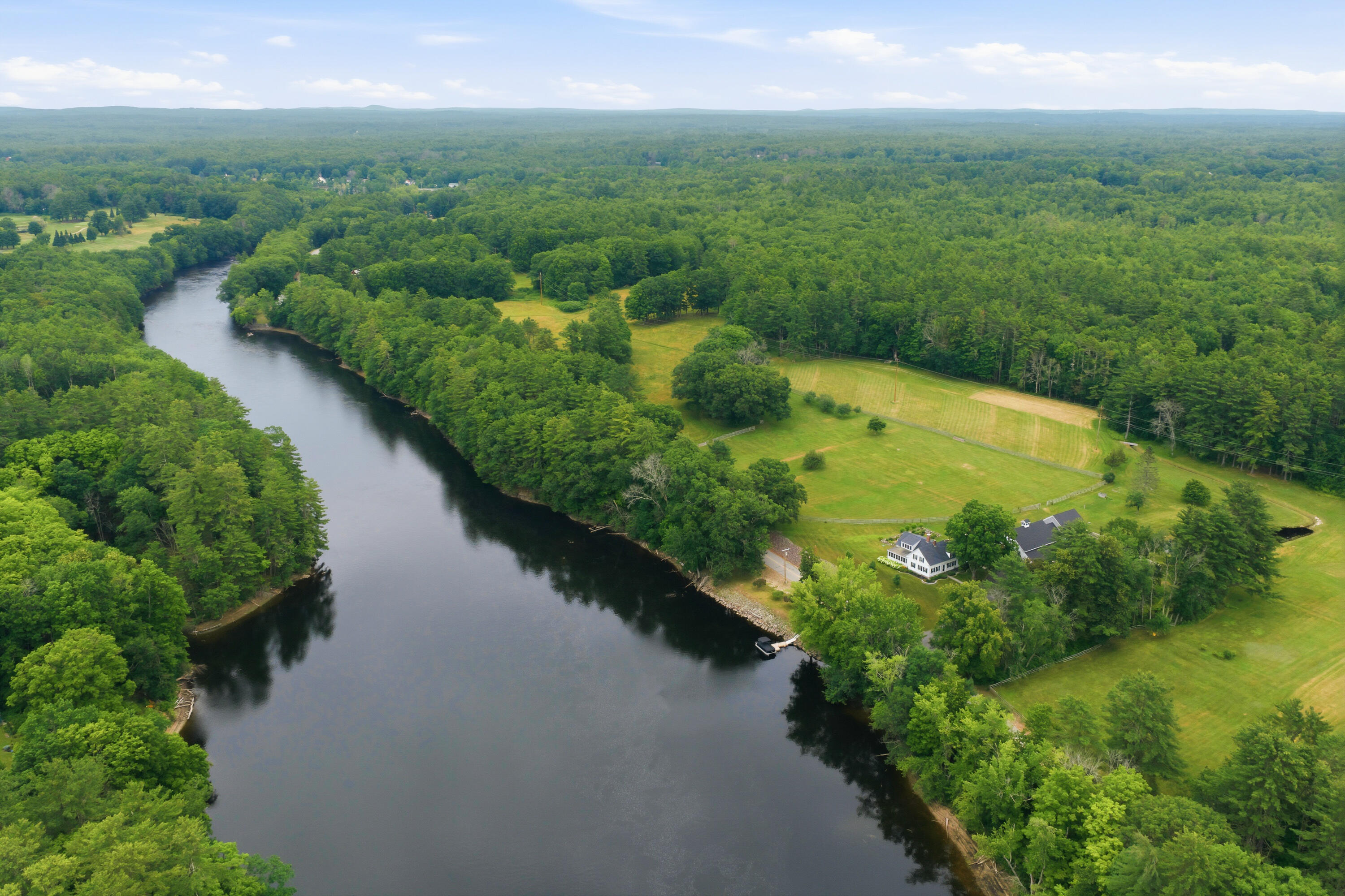 Aerial view of a riverside home with a dock, open lawns, and surrounding woodland.