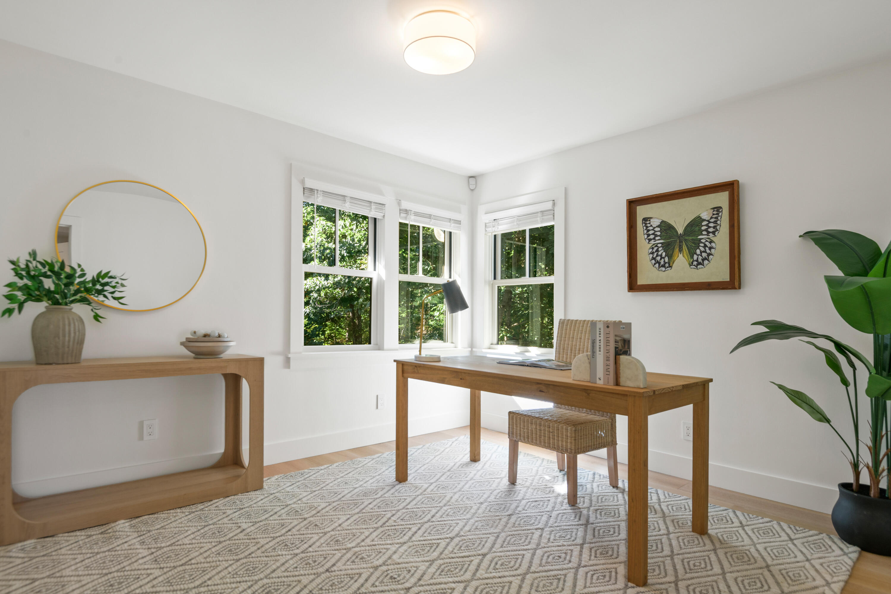 Bright home office with a wooden desk, wicker stool, and windows overlooking leafy greenery.