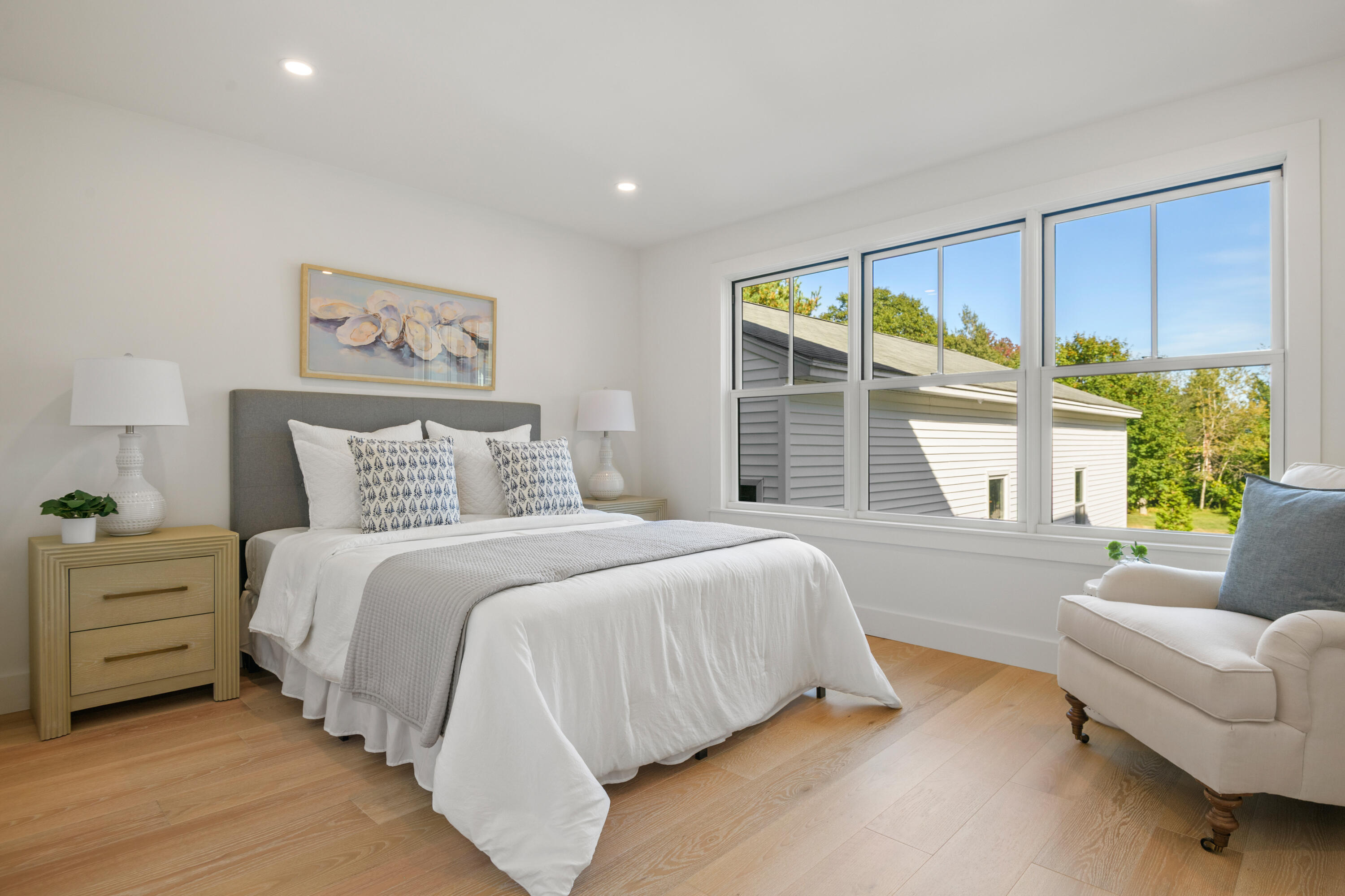 Staged bedroom with a white bed, gray headboard, and large windows overlooking trees.