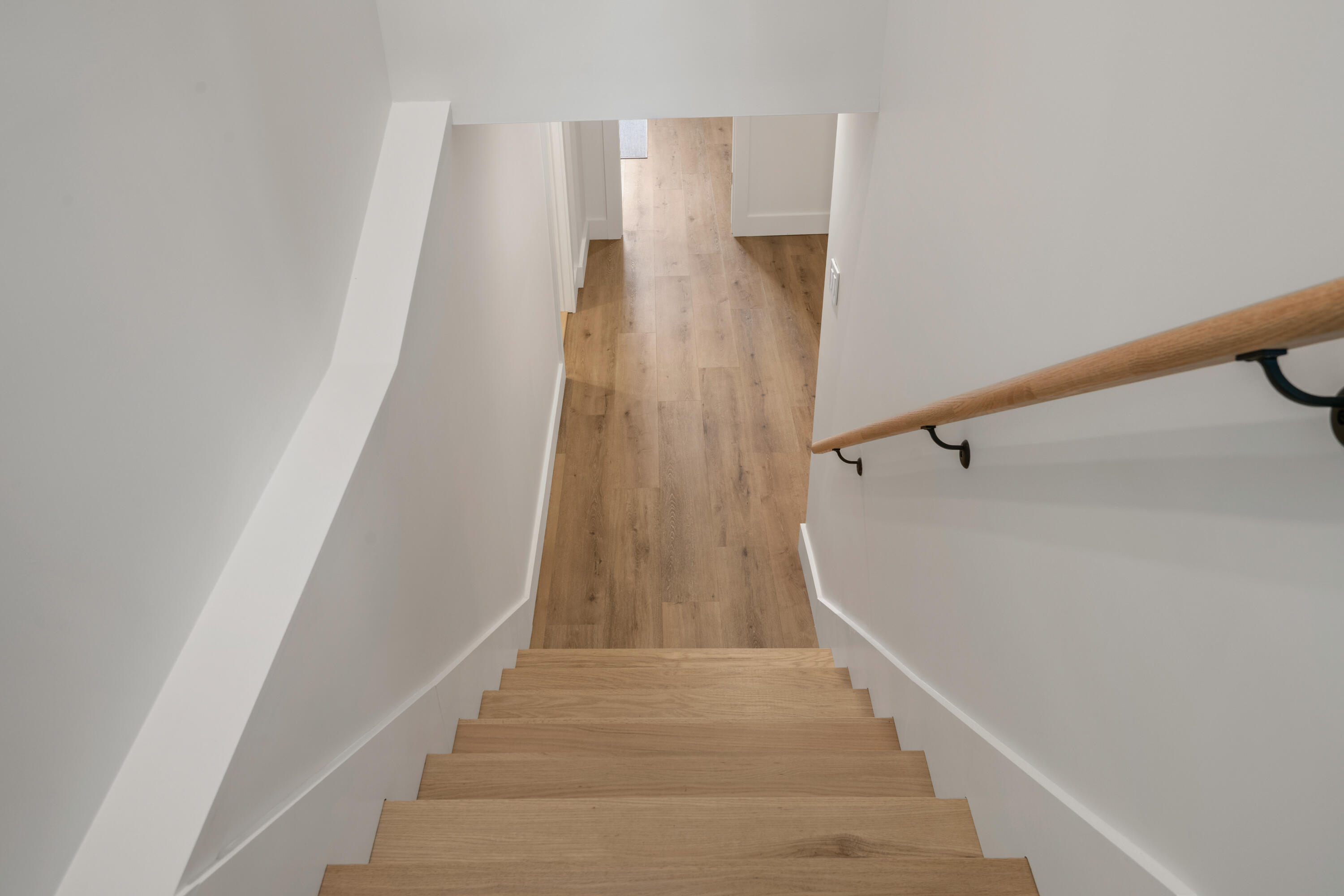 Light wood staircase descends to a hallway with plank flooring and a wooden handrail.