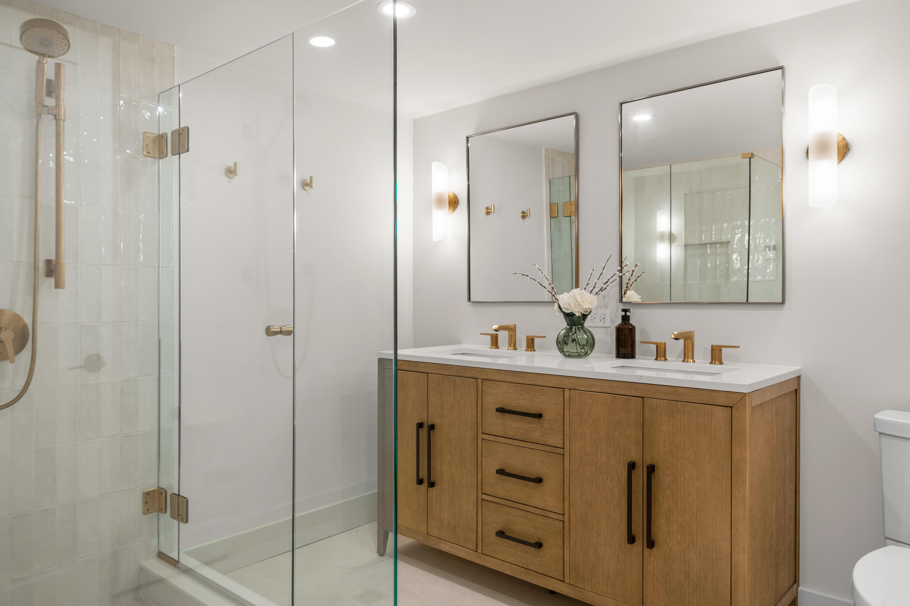 Modern bathroom with a glass shower and wood double vanity featuring brass fixtures.